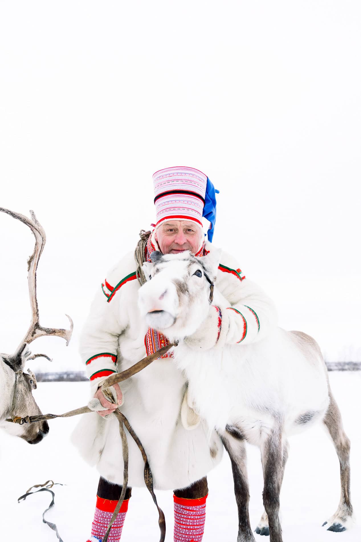 Sami man in traditional clothing with reindeer in snowy landscape.